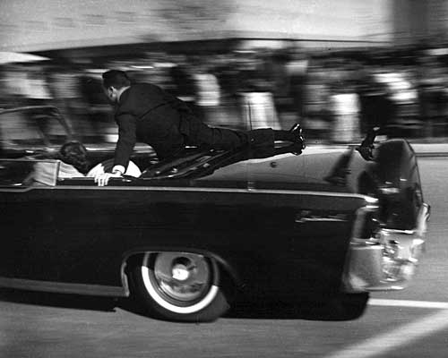 The presidential limousine, a 1961 Lincoln Continental, races toward Parkland Hospital, with Secret Service Agent Clinton Hill clinging to the back.